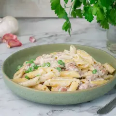 En krämig kyckling pasta med ärtor i en grön skål, med vitlök och persilja i bakgrunden.