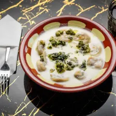 En skål med shish barak i en krämig sås, toppad med örter. Bredvid en läsk och bestick på ett svart bord med gulddetaljer.