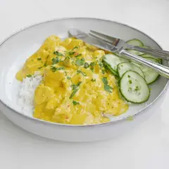 En skål med ris och krämig kycklingcurry, toppad med persilja. Gurkskivor och bestick ligger bredvid. Curry nam nam!