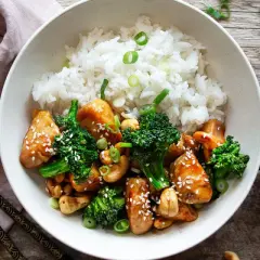 En skål med ris, kyckling och broccoli, toppad med sesamfrön och salladslök. Bredvid syns en flaska kycklingfond.