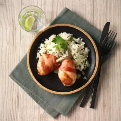 En tallrik med kycklingklubbor inslagna i bacon och krämig risotto. Ett glas vatten med lime och bestick ligger bredvid. Ett perfekt risotto recept kyckling!
