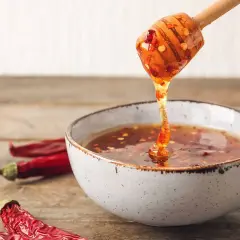 En träsked droppar gyllene chili honung ner i en skål. Torkade chilifrukter ligger bredvid.