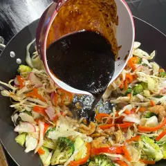 Mörk, glansig wok sås hälls över färgglada grönsaker som broccoli, paprika och kål i en het wokpanna.