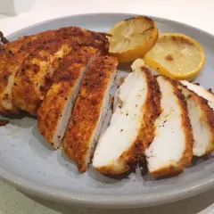 Skivad panerad kyckling med kryddig yta, serverad med grillade citronklyftor på en grå tallrik.