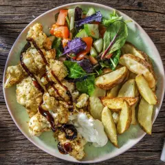 En tallrik med rostad blomkål i ugn, klyftpotatis och en fräsch sallad. Blomkålen är täckt med ströbröd och ringlad med en mörk sås.