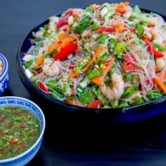 En stor skål med glasnudel sallad med räkor, paprika och grönsaker. Bredvid finns en skål med dressing och en skål med hackade nötter.
