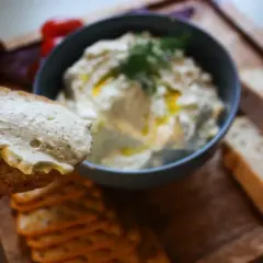 En bit bröd med krämig vispad fetaost och dill. I bakgrunden en skål med mer ostdipp och kex.
