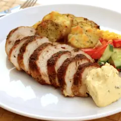 En tallrik med saftig kycklingfilé, skivad och kryddad, serverad med ugnsrostade potatisskivor, majs, paprika och gurka, samt en klick krämig sås.