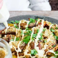 En hand spritsar ponzu mayo över krispiga potatisklyftor, ströbröd och färsk persilja på en mörk tallrik.