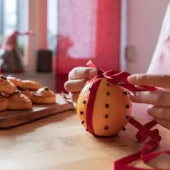 Händer knyter ett rött band runt en apelsin med nejlika hel. Bredvid ligger saffransbullar.