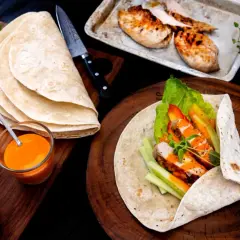 En färdig kyckling wrap med sallad, paprika och gurka. Bredvid ligger tortillabröd, dressing och grillad kyckling.