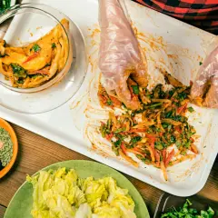 Händer i plasthandskar blandar ingredienser för ett kimchi recept. Grönsaker och kryddor ligger upplagda runt om.