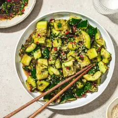 En skål med smashad gurksallad, toppad med sesamfrön och örter, med ätpinnar vilande ovanpå.