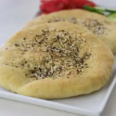 Två runda manakish zaatar med sesamfrön och örter på en vit bricka, med skivad gurka och tomat i bakgrunden.