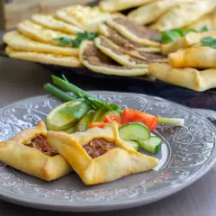 Två lahm bi ajin, små köttfyllda bröd, serveras på en grå tallrik med grönsaker. I bakgrunden syns fler lahm bi ajin och andra bakverk.