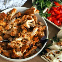 En skål med kryddad kyckling för kyckling gyros, bredvid grönsaker och pitabröd.