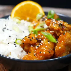 En skål med ris och krispig, glaserad orange chicken, toppad med sesamfrön och salladslök. En apelsinskiva vilar bakom.