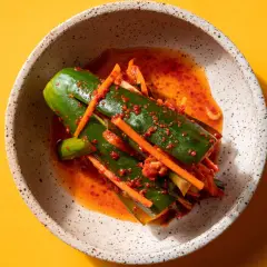 En skål med kimchi gurka, kryddad med chili och morot, på en gul bakgrund.