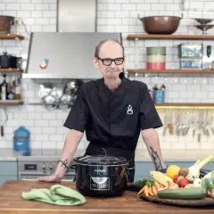 En kock med tatueringar står vid ett köksbord med en slow cooker och färska grönsaker. Perfekt för att hitta nya ångkokare recept.