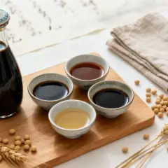 En flaska mörk sojasås och skålar med olika sorters sojasås, omgivna av sojabönor och veteax. Sojasås görs av fermenterade sojabönor.