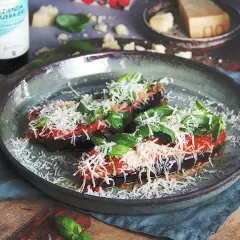 Två halvor av aubergine i ugn, toppade med tomatsås, basilika och riven ost. Serveras på en rustik tallrik.