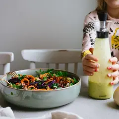 En skål med färgglad sallad och två flaskor dressing, en grön och en ljusgul. En flicka håller i den gröna flaskan.