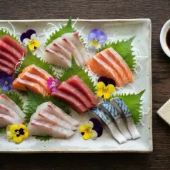 Vacker sashimi-tallrik med olika fiskar, garnerad med blommor. Bredvid finns en skål med sushi sås och en färsk wasabirot.
