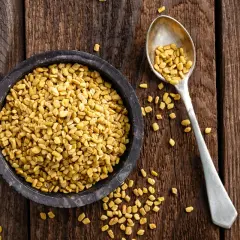 En skål med gyllene bockhornsklöverfrön (fenugreek seeds svenska) bredvid en sked full med samma frön, på en rustik träbakgrund.