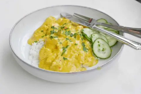 En skål med ris och krämig kycklingcurry, toppad med persilja. Gurkskivor och bestick ligger bredvid. Curry nam nam!