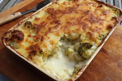 En ugnsform med ostgratinerad blomkål och broccoli, gyllene och bubblande. En sked ligger bredvid.