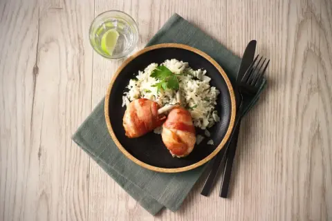 En tallrik med kycklingklubbor inslagna i bacon och krämig risotto. Ett glas vatten med lime och bestick ligger bredvid. Ett perfekt risotto recept kyckling!