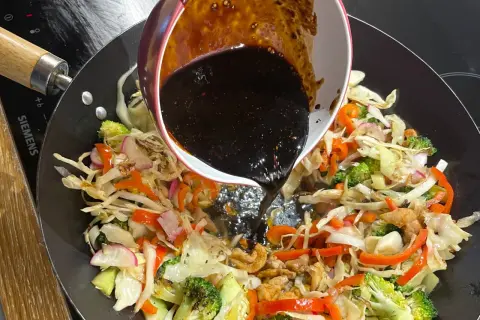 Mörk, glansig wok sås hälls över färgglada grönsaker som broccoli, paprika och kål i en het wokpanna.