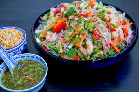 En stor skål med glasnudel sallad med räkor, paprika och grönsaker. Bredvid finns en skål med dressing och en skål med hackade nötter.