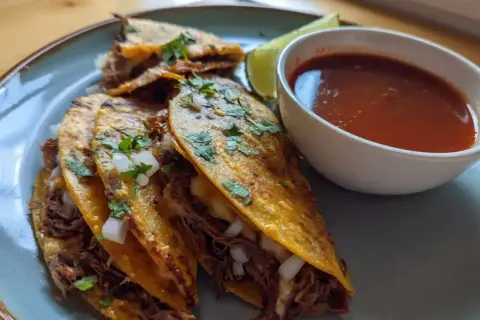 Tre saftiga birria tacos med strimlat kött, lök och koriander, serverade med en skål consommé och en limeklyfta.