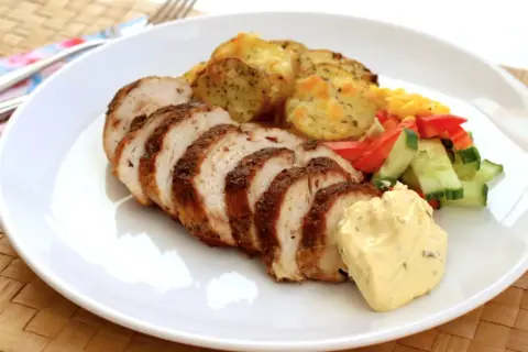 En tallrik med saftig kycklingfilé, skivad och kryddad, serverad med ugnsrostade potatisskivor, majs, paprika och gurka, samt en klick krämig sås.