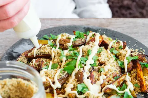 En hand spritsar ponzu mayo över krispiga potatisklyftor, ströbröd och färsk persilja på en mörk tallrik.