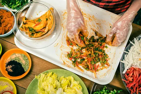 Händer i plasthandskar blandar ingredienser för ett kimchi recept. Grönsaker och kryddor ligger upplagda runt om.
