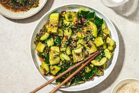 En skål med smashad gurksallad, toppad med sesamfrön och örter, med ätpinnar vilande ovanpå.