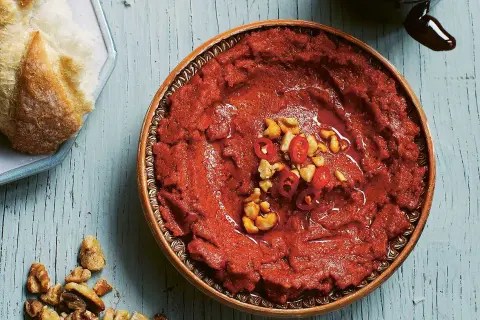 En skål med röd muhammara, toppad med chili och nötter, bredvid en bit bröd. Perfekt för ditt muhammara recept.