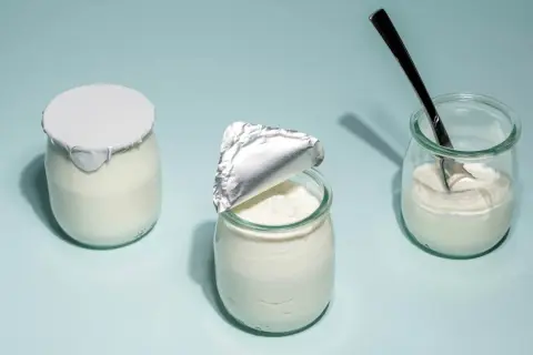 Tre burkar med hemmagjord yoghurt på ljusblå bakgrund. En är helt försluten, en annan är delvis öppnad och en tredje har en sked i sig.