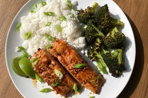 Två filéer av asiatisk lax med sesamfrön, serveras med ris, rostad broccoli och limeklyftor.