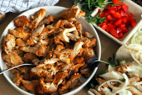 En skål med kryddad kyckling för kyckling gyros, bredvid grönsaker och pitabröd.