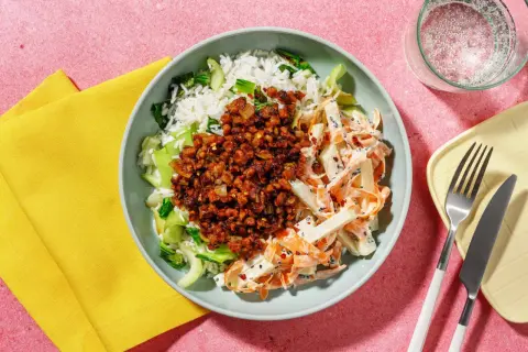 En skål med ris, pak choi sallad, och en kryddig topping. Bredvid ligger en gul servett och bestick.