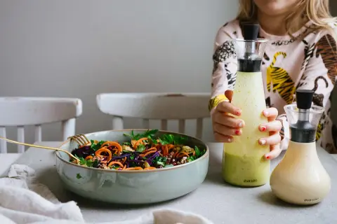 En skål med färgglad sallad och två flaskor dressing, en grön och en ljusgul. En flicka håller i den gröna flaskan.
