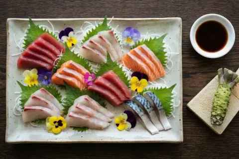 Vacker sashimi-tallrik med olika fiskar, garnerad med blommor. Bredvid finns en skål med sushi sås och en färsk wasabirot.