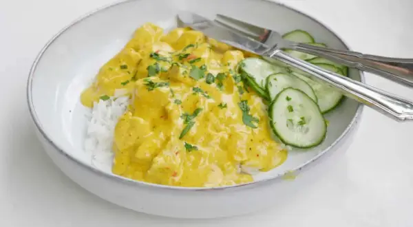 En skål med ris och krämig kycklingcurry, toppad med persilja. Gurkskivor och bestick ligger bredvid. Curry nam nam!