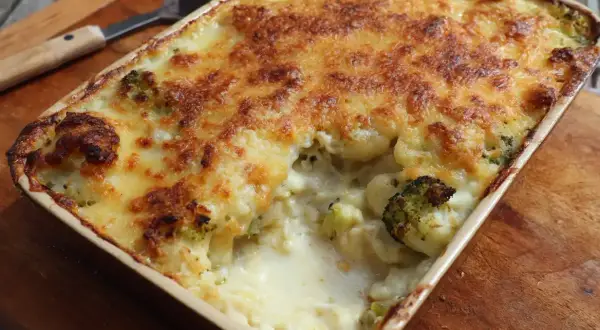 En ugnsform med ostgratinerad blomkål och broccoli, gyllene och bubblande. En sked ligger bredvid.