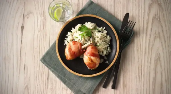 En tallrik med kycklingklubbor inslagna i bacon och krämig risotto. Ett glas vatten med lime och bestick ligger bredvid. Ett perfekt risotto recept kyckling!