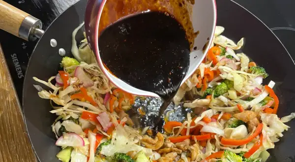 Mörk, glansig wok sås hälls över färgglada grönsaker som broccoli, paprika och kål i en het wokpanna.
