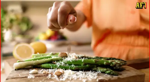 En hand strör parmesan över sparris som förbereds för att grilla. Citroner och nötter syns i bakgrunden.